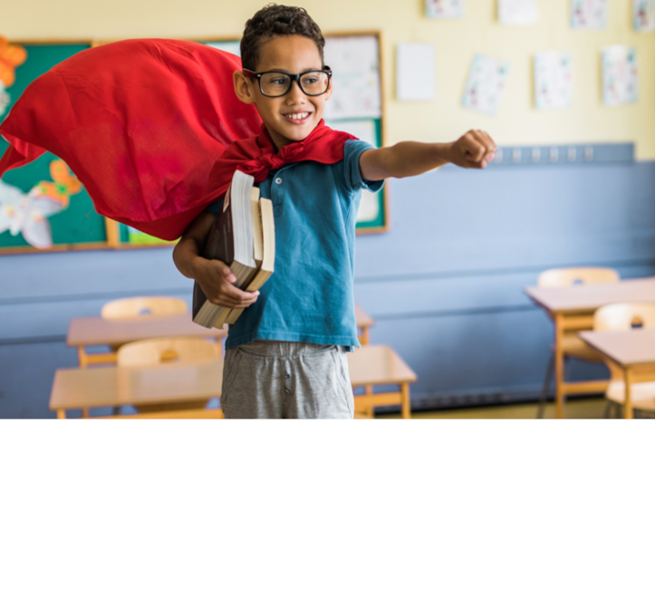 Junge im Heldenkostüm in Klassenzimmer mit Büchern unter dem Arm